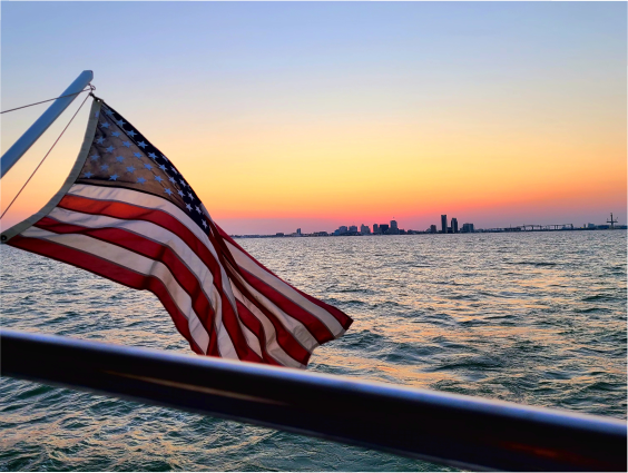 United States flag waving on a boat at sunset with Corpus Christi city skyline visible across the water in the distance