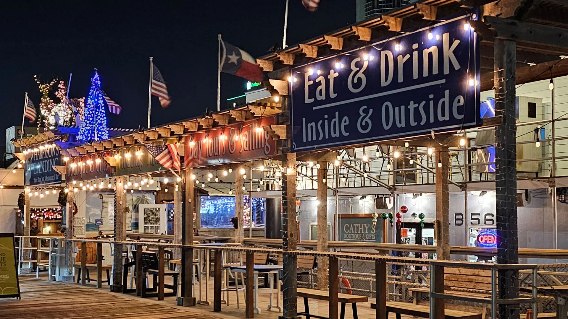 Harrison's Landing restaurant, Boutique & Gifts, Tiki Bar, and Bay Tours with vibrant signs and flags inviting patrons to eat and drink inside or outside at night