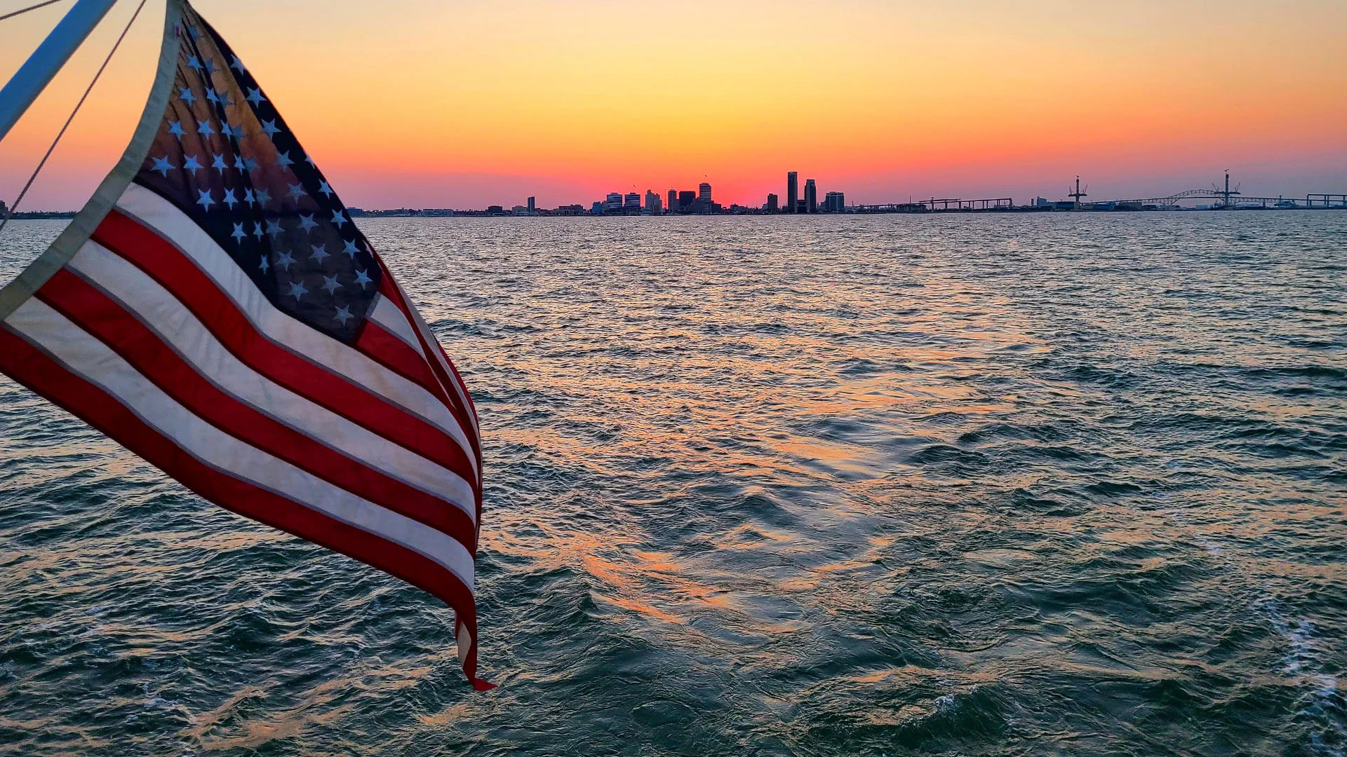 United States flag waving on a boat at sunset with Corpus Christi city skyline visible across the water in the distance